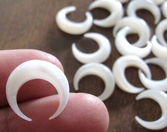 May include: A collection of white, crescent-shaped jewelry components. One is held between fingers, showcasing its smooth, pearly surface. The other components are scattered on a brown surface, suggesting a craft or jewelry-making theme.