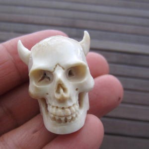May include: A carved skull figurine with small horns, crafted from a light-colored material. The skull features detailed eye sockets, a nose cavity, and a full set of teeth. The horns curve slightly upward. The figurine is held in a hand.