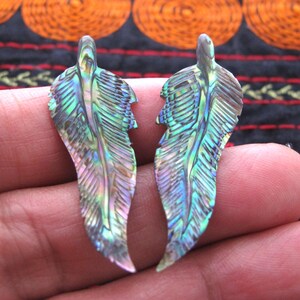 May include: Two iridescent abalone shell feather pendants. The pendants display a spectrum of colours, including green, blue, and purple, with intricate veining details. The feathers are held in a person's hand.