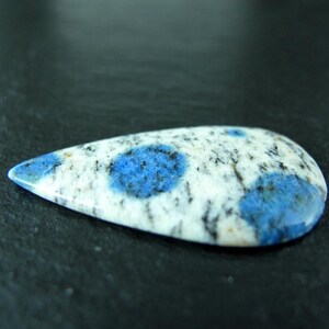 May include: A teardrop-shaped cabochon of blue and white granite with a polished surface. The stone has a speckled pattern of blue and white.