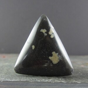 May include: A triangular cabochon of black obsidian with white inclusions. The stone is polished and has a smooth, glossy finish.