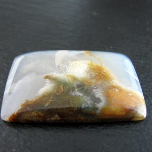 May include: A white agate cabochon with brown and green inclusions. The stone is translucent and has a smooth, polished surface.