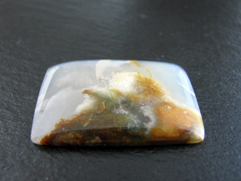 May include: A white agate cabochon with brown and green inclusions. The stone is translucent and has a smooth, polished surface.