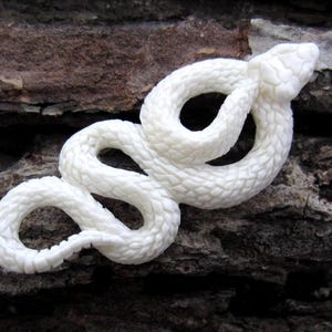 May include: A coiled, white snake sculpture with detailed scales. The snake is carved from a solid material and rests on a textured, brown surface. The snake's head is raised, giving it a lifelike appearance.