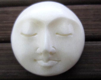 May include: A round, off-white sculpture of a serene face with closed eyes and a gentle smile. The smooth, rounded form suggests a celestial theme, possibly representing the moon or a similar figure.