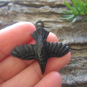 May include: Black wooden bird pendant with detailed carving. The bird is in flight with its wings spread out. The pendant has a loop at the top for attaching a chain or cord.