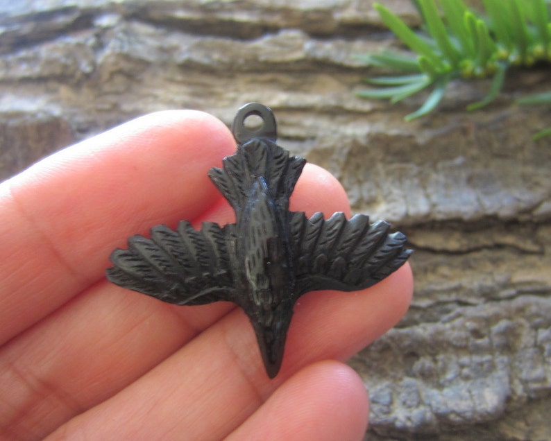 May include: Black wooden bird pendant with detailed carving. The bird is in flight with its wings spread out. The pendant has a loop at the top for attaching a chain or cord.