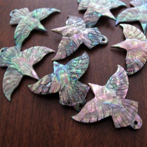May include: A collection of iridescent bird-shaped pendants. Each pendant is carved with feather details and has a small hole for stringing. The pendants display a range of colors, including green, blue, and purple, and are set against a dark brown background.