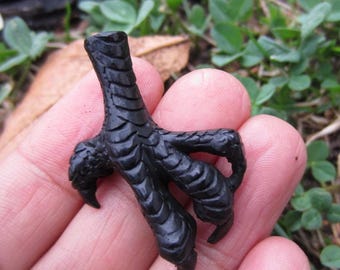 Puede incluir: Un colgante de garra de ave en color negro mate que presenta escamas texturizadas detalladas a lo largo de la pata y garras afiladas y curvas. El objeto está esculpido con una estructura anatómica realista, un acabado oscuro y una abertura hueca.