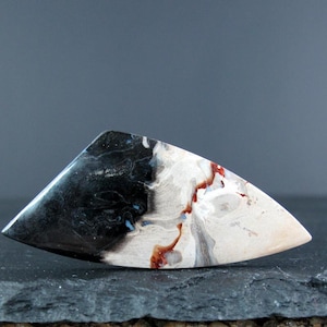 May include: A white, black, and red agate cabochon with a unique, abstract pattern. The cabochon is shaped like a teardrop and has a smooth, polished surface.