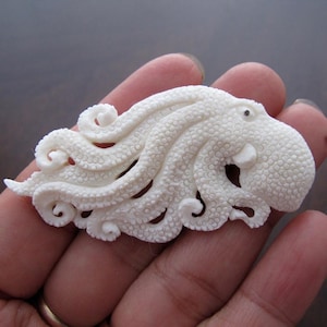 May include: A detailed, off-white octopus sculpture with textured skin and curled tentacles. The sculpture has a small, dark eye and is carved with intricate detail, showcasing the form of the sea creature. The octopus is a decorative item.