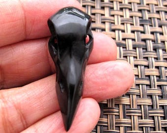 Puede incluir: Colgante de calavera de cuervo negro brillante. La calavera tallada presenta un pico puntiagudo y cuencas de los ojos detalladas. El colgante se sostiene en una mano sobre un fondo tejido. Un accesorio de estilo gótico o alternativo.
