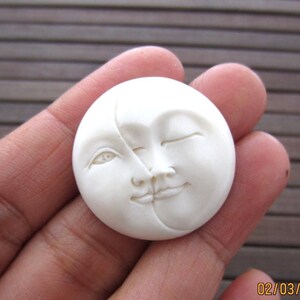 May include: A white, circular carving of a smiling moon face. The moon is depicted with one eye open and the other closed, with detailed facial features. The carving is held in a hand.