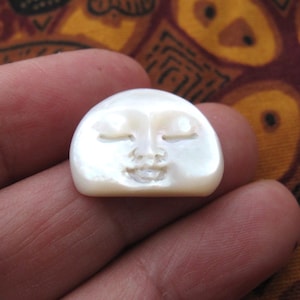 Può includere: Un piccolo volto di luna scolpito, bianco sporco, con gli occhi chiusi e la bocca sorridente. Il volto lunare è liscio e arrotondato, con una lucentezza perlacea. L'intaglio è dettagliato e delicato, con ombre sottili.