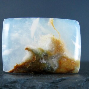 May include: A white, translucent cabochon with brown and green inclusions. The stone has a cloudy, agate-like appearance.