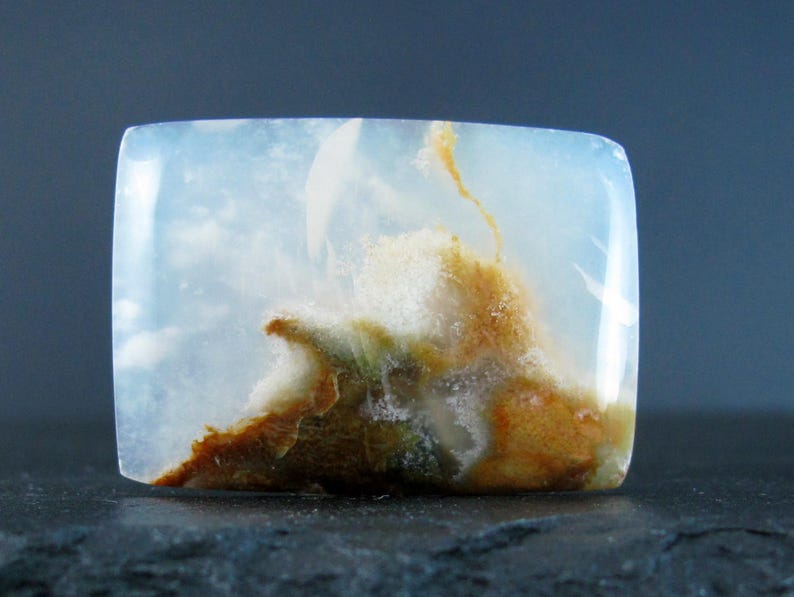 May include: A white, translucent cabochon with brown and green inclusions. The stone has a cloudy, agate-like appearance.