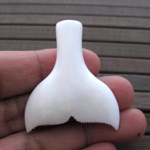 May include: A white, whale tail-shaped bottle stopper. The stopper is smooth and glossy, with a rounded top and a tail fin design. The tail fin has a scalloped edge.