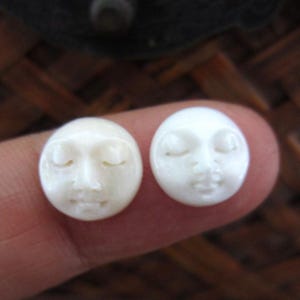 May include: Two small, oval-shaped, carved faces with closed eyes. One is a creamy off-white color, and the other is a bright white. The faces have delicate features, including a nose and a smiling mouth. The faces are held by a finger.