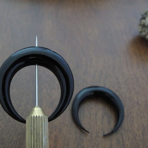K&ouml;nnte beinhalten: Zwei schwarze Horn-Halbmond-Ohrringe. Die Ohrringe werden auf einer Holzoberfl&auml;che gezeigt. Ein Ohrring wird von einem Werkzeug mit einer scharfen Spitze gehalten.