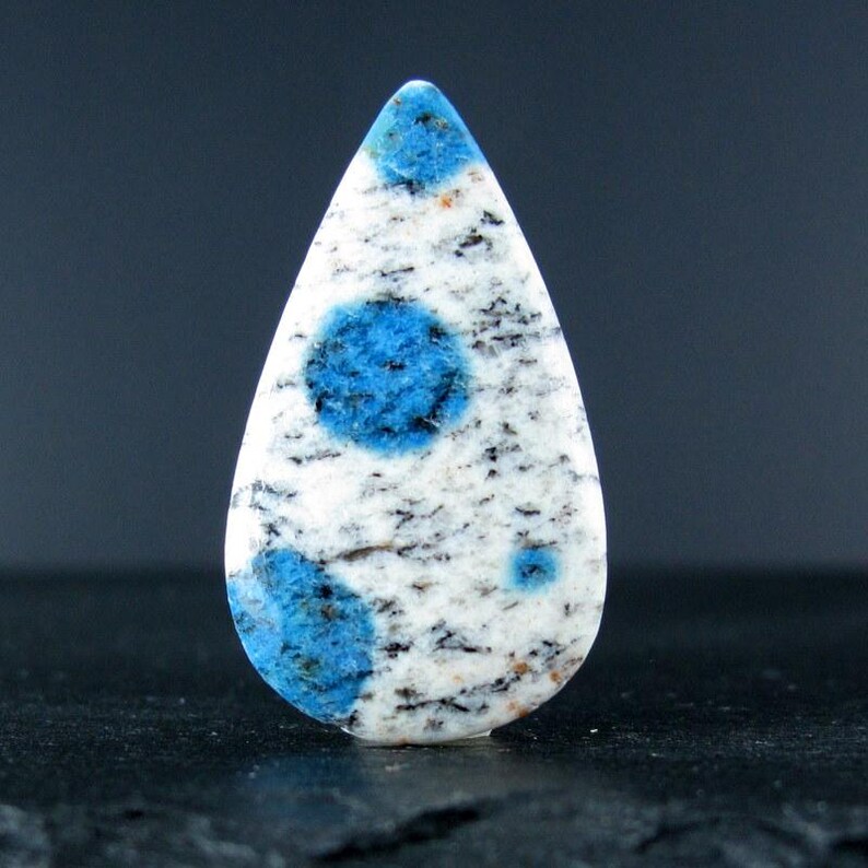 May include: A teardrop-shaped cabochon of blue and white kyanite, a type of gemstone. The stone has a white background with blue spots.