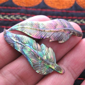 May include: Two iridescent feather-shaped pendants. The pendants display a spectrum of colours, including green, purple, and gold, with intricate carved details. The feathers are held in a hand, showcasing their natural beauty and craftsmanship. These pendants are ideal for jewellery making.