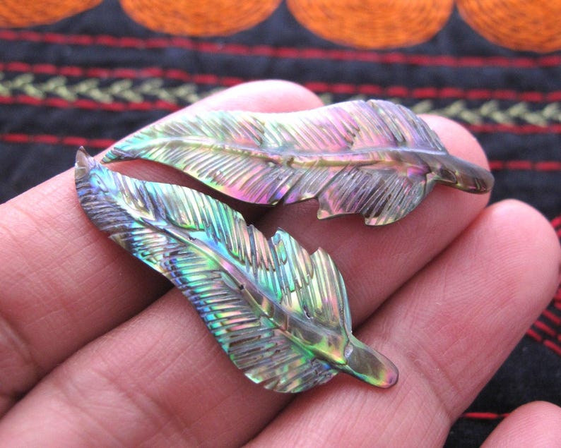 May include: Two iridescent feather-shaped pendants. The pendants display a spectrum of colours, including green, purple, and gold, with intricate carved details. The feathers are held in a hand, showcasing their natural beauty and craftsmanship. These pendants are ideal for jewellery making.