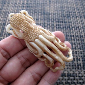 May include: A detailed, cream-colored octopus carving with a textured body and curled tentacles. The octopus is held in a hand, showcasing the intricate details of the sculpture. The carving has a light brown hue.