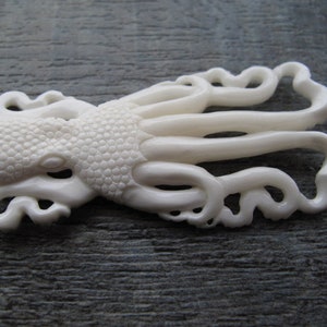May include: White carved octopus pendant made of bone. The octopus has detailed tentacles and a textured body.
