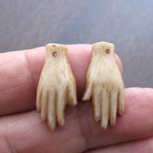 May include: Two small, cream-colored hand-shaped charms. Each charm has detailed fingers and a small hole at the top. The charms have a slightly aged appearance, with subtle brown shading.