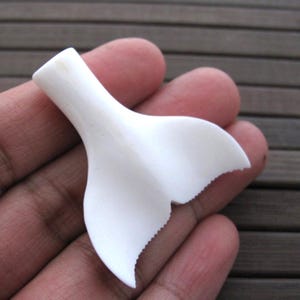 May include: A white, carved whale tail pendant. The pendant is held in a hand and has a smooth, curved design with a detailed, scalloped edge. The pendant is made of a solid, opaque material.