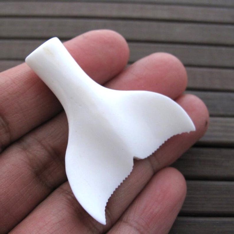 May include: A white, carved whale tail pendant. The pendant is held in a hand and has a smooth, curved design with a detailed, scalloped edge. The pendant is made of a solid, opaque material.