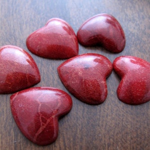 May include: Six red heart-shaped coral stones on a brown wooden surface.