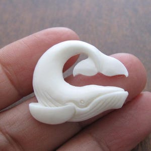 May include: A white whale carved from bone, with a detailed design of the whale's body and tail. The whale is carved in a leaping position, with its tail raised in the air.