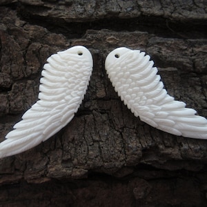 May include: Two white carved bone angel wings with detailed feather patterns. The wings are symmetrical and have a hole at the top for hanging.