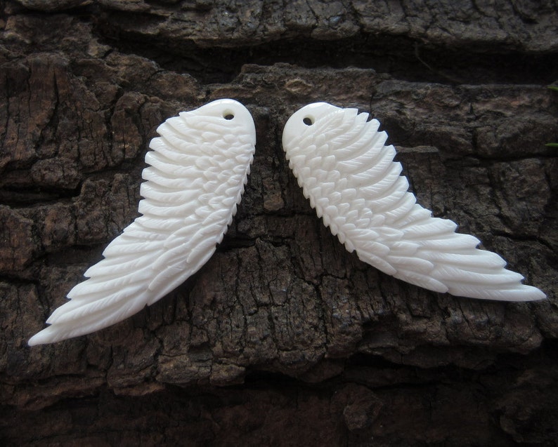 Puede incluir: Dos alas de &aacute;ngel blancas talladas en hueso con patrones de plumas detallados. Las alas son sim&eacute;tricas y tienen un agujero en la parte superior para colgar.