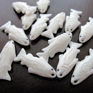 May include: A collection of small, white, fish-shaped carvings. Each carving has detailed fins, scales, and a small black dot for an eye. The carvings are scattered on a dark surface, creating a textured and artistic display.