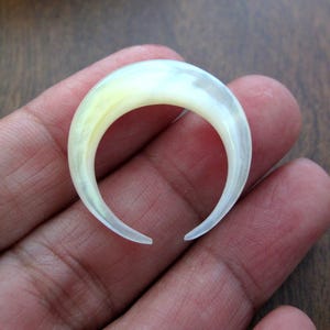 May include: A crescent-shaped, off-white ear gauge made of a translucent material. The gauge has a smooth, polished surface with subtle yellow and gray hues. The open ends of the crescent are rounded.