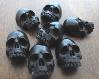 May include: A collection of small, glossy black skull beads. Each skull has detailed facial features, including eye sockets, a nose cavity, and teeth. The skulls are arranged on a wooden surface, creating a gothic or Halloween theme.