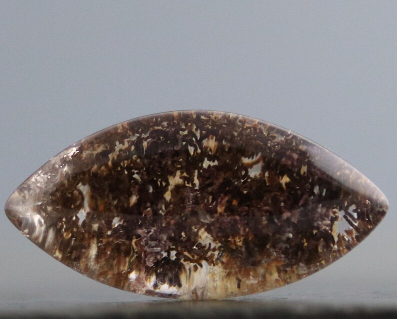 May include: A clear, faceted, teardrop-shaped gemstone with brown and white dendritic inclusions. The gemstone has a smooth, polished surface.