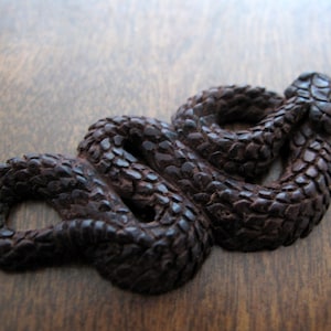 Puede incluir: Una escultura de serpiente de madera marrón oscuro con una superficie texturizada. La serpiente está enroscada en forma de ocho.