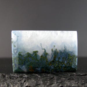 May include: A rectangular moss agate gemstone with a landscape design. The top portion is white and translucent, transitioning to a blue and green landscape at the bottom. The stone is set against a dark background.