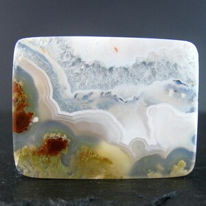 May include: A polished agate cabochon with a white, grey, and brown banded pattern. The agate has a natural, organic look with a variety of colours and textures.
