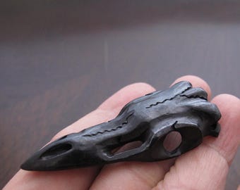 Puede incluir: Un cráneo de cuervo tallado en gris oscuro. El cráneo se sostiene en una mano y presenta tallas detalladas, incluyendo las cuencas de los ojos y el pico. El cráneo de cuervo es un artículo decorativo único.