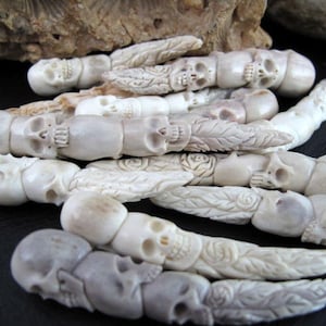 May include: Close-up of several carved bone skull beads. The beads are strung together, forming a chain-like structure. Some beads have floral or leaf designs. The color is off-white with some variations in tone.