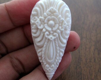May include: A teardrop-shaped white pendant featuring an intricate, raised floral and scrollwork relief carving. The surface displays a symmetrical pattern with a central flower motif surrounded by detailed, flowing lines and decorative borders.