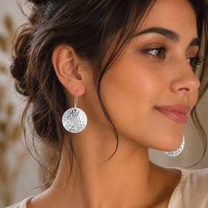May include: A pair of silver-toned drop earrings featuring circular discs with a hammered, textured finish. The earrings are suspended from thin, curved metal hooks that pass through the earlobe, creating a dangling effect with a reflective surface.