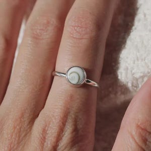 Può includere: Un anello in argento con una montatura circolare contenente un inserto di conchiglia a spirale bianca e marrone. La fascia è sottile e lucida, e trattiene il pezzo di conchiglia rotondo in modo sicuro all'interno di una montatura a castone.
