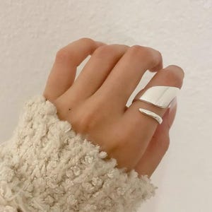 May include: A polished silver ring featuring an open, bypass-style design with a wide, flat, curved upper band and a thinner, rounded lower band that wraps around the finger, creating a sleek, reflective surface with a smooth, metallic finish.