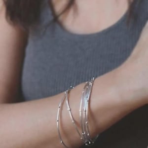 May include: A set of three thin, silver-toned metal bangles worn on the wrist. Each bangle features a smooth, circular wire design accented with small, evenly spaced spherical metal beads along the entire circumference of the band.
