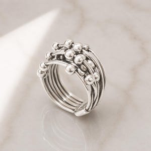 Könnte beinhalten: Ein silberfarbener Ring bestehend aus mehreren dünnen, übereinanderliegenden Bändern. Die Außenfläche des Rings ist mit einer Reihe kleiner, polierter, kugelförmiger Perlen verziert, die an den einzelnen drahtartigen Bändern befestigt sind.